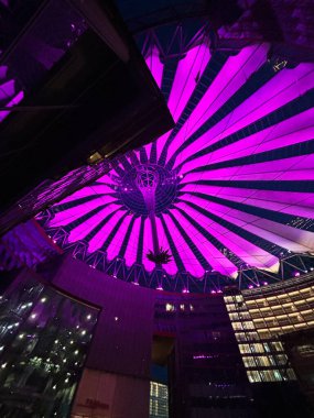 Berlin 'deki canlı bir mor tepe örtüsü Sony Center modern kentsel mimarinin üstündeki gece gökyüzünü aydınlatır. Karmaşık geometrik tasarım büyüleyici bir görsel etki yaratır..