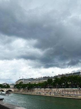 Paris 'te bulutlu bir gün ve nehir boyunca klasik mimari manzarası var. Sahne, dramatik bir gökyüzü altında şehrin sakin atmosferini yakalıyor..