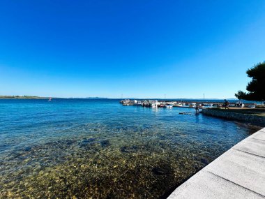 Berrak mavi suların, taş bir iskelenin ve dalgalı bir gökyüzünün altına yanaşmış teknelerin bulunduğu pitoresk bir sahil manzarası. Huzuru ve tatil beldelerini göstermek için ideal.