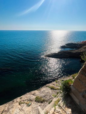 Güneşli, kayalık bir sahil şeridinin berrak mavi gökyüzünün altında parıldayan deniz manzarası. Sakin sahne huzuru ve doğal güzelliği çağrıştırıyor. Ulcinj, Karadağ