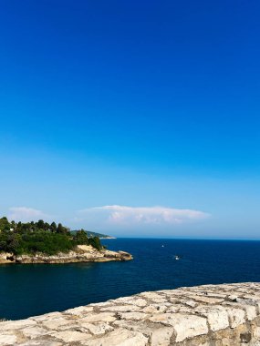 Dalgalı mavi bir denize karşı kayalık bir uçurum ve yemyeşil bir yeşillik barındıran çarpıcı bir kıyı manzarası. Açık gökyüzü, okyanus manzarasının sükunetini ve doğal güzelliğini arttırır. Ulcinj, Karadağ