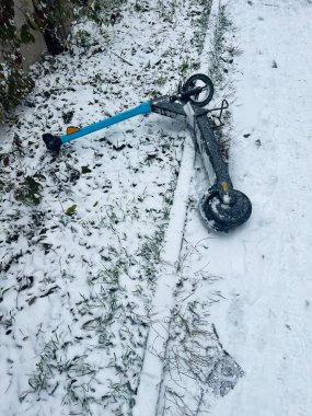 An electric scooter lies abandoned on a snow-covered path, surrounded by grass and trees. The image captures a sense of neglect and wintery chill. clipart