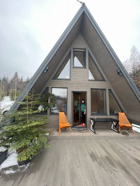 Huzurlu bir karlı ormanda, turuncu koltukları ve tatil kaçamakları ve rahatlama için mükemmel neşeli bir atmosferi olan hoş bir açık hava güvertesi olan büyüleyici bir A-frame kabin..