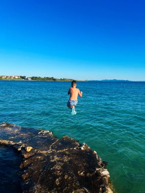 Genç bir çocuk bulutsuz gökyüzünün altındaki kayalık bir sahilden kristal berrak mavi denize atlıyor, neşe ve macerayı somutlaştırıyor. Kıyı manzarası arka planda görünür..