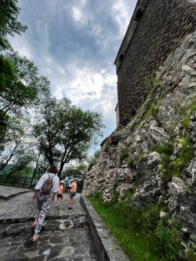 Bir grup insan, tarihi bir taş duvarın yanında, yeşil ve bulutlu bir gökyüzünün altında, taştan bir yolda yürüyor. Bran, Romanya.