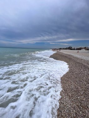 Sakin bir kıyı manzarası hafif dalgaların dramatik bulutlu bir gökyüzünün altında çakıllı bir sahile çarptığını gösteriyor. Rahatlama, doğa ve sükunet temaları için ideal. Porto Sant 'Elpidio, İtalya.