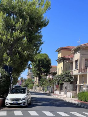Porto Sant 'Elpidio, İtalya - 6 Temmuz 2022: Açık mavi gökyüzü altında park halindeki beyaz bir araba, canlı bir yeşillik ve büyüleyici konut evleri içeren sakin bir banliyö caddesi.