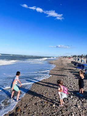 Üç çocuk kumsalda neşeli bir günün tadını çıkarıyorlar, dalgaları hissediyorlar ve açık mavi bir gökyüzünün altında kıyıları keşfediyorlar. Porto Sant 'Elpidio, İtalya.
