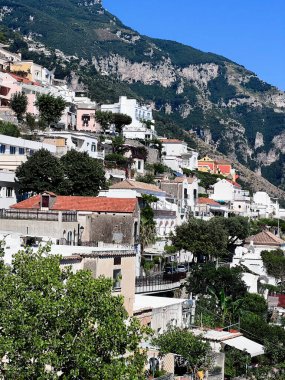 Berrak mavi gökyüzünün altında yemyeşil yemyeşil ortamda renkli evlerin yer aldığı pitoresk tepe kenarı köyü. Huzurlu ve manzaralı manzara, Akdeniz mimarisinin cazibesini yansıtıyor. Pozitano, İtalya.