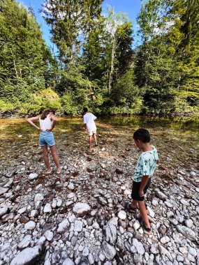 Üç çocuk, bir orman nehrinin kenarında kayalık nehir yatağını keşfederken güneşli bir günün tadını çıkarırken görülüyor. Bereketli yeşillik ve berrak gökyüzü açık havayı güzelleştirir. Bohinj, Slovenya.