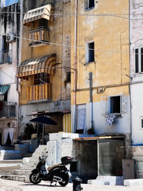 Balkonları ve çizgili tenteleri olan yıpranmış sarı bir binanın park etmiş bir scooterla eşlik ettiği büyüleyici, kırsal bir şehir manzarası. Macera ve Akdeniz yaşam tarzları. Bari, İtalya.