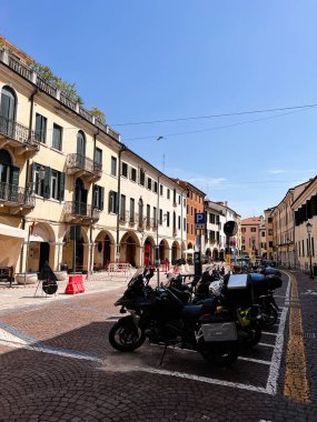 Padua, İtalya - 4 Temmuz 2022: Tarihi mimari ve park edilmiş motosikletlerin sergilendiği bir İtalyan caddesi görüntüsü. Sahne, şehir hayatının cazibesini parlak mavi bir gökyüzünün altında yakalıyor.