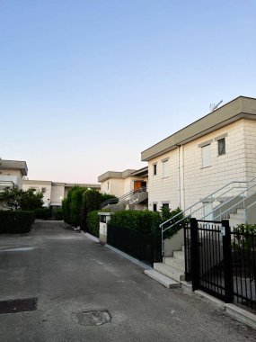 A quiet residential street lined with contemporary houses under a clear blue sky. The peaceful atmosphere is enhanced by well-maintained greenery and evening lighting. Ippocampo, Italy. clipart