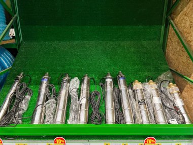 Kyiv, Ukraine - January 03, 2025: A shelf in a store displaying multiple stainless steel submersible pumps with wires and labels, set against green carpet-like background. This image illustrates hydraulic equipment and technology for water systems. clipart