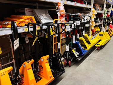 A selection of industrial pallet trucks in various colors and sizes in a retail store aisle, with other tools and equipment displayed on surrounding shelves for purchase. clipart