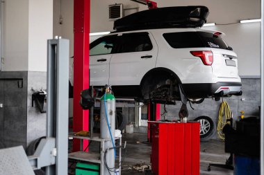 Otomobil tamirhanesinde beyaz bir SUV hidrolik asansörde yükselir. Sahne, çeşitli araç ve gereçlerle modern ve iyi donanımlı bir atölye ortamını gösteriyor..
