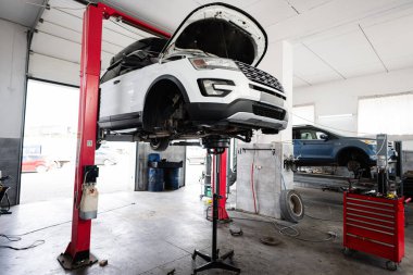 Profesyonel bir oto tamirhanesinde bir araç bakım görüyor. Beyaz bir araba hidrolik asansörle yükseltilir, atölye ortamını ve araçları gösterir.