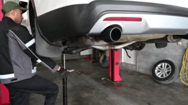 Üniformalı bir tamirci kaldırılmış bir arabanın altında çalışıyor. Aracı desteklemek için kriko kullanıyor..