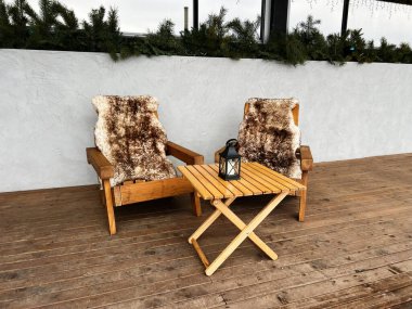 A wooden seating area featuring two chairs with fur covers, a small table, and a lantern. Perfect for a rustic, comfortable ambiance on a porch or patio. clipart