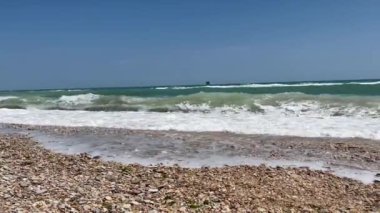 Güneşli bir günde dalgalar çakıl taşı plajına çarpıyor. Turkuaz su berrak mavi gökyüzünün altında parıldıyor..