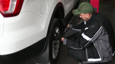 Üniformalı profesyonel bir tamirci, iyi donanımlı bir oto tamirhanesinde beyaz bir cipin lastiğini değiştiriyor. Bu video araba bakım, onarım ve kalifiye işçilik temaları çağrıştırıyor.