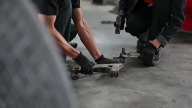 Garajdaki iki tamirci, çekiç ve ingiliz anahtarı kullanarak paslı bir araba parçasını çıkarmak için birlikte çalışıyorlar..