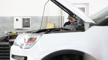 Üniformalı bir tamirci, iyi aydınlatılmış bir tamirhanede beyaz bir SUV 'nin motorunu özenle inceliyor..