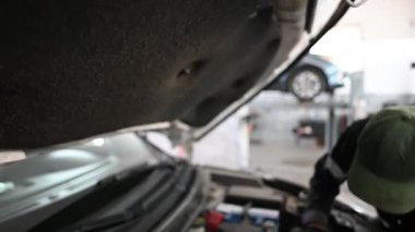 Bir tamirci bir araba motoru üzerinde çalışıyor. O, siyah bir üniforma ve yeşil bir şapka giyiyor. Araba asansörde ve kaput açık. Araçlar ve parçalar görünür.