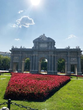 Madrid, İspanya - 22 Ağustos 2024: Madrid, İspanya 'da güneşli bir gökyüzünün altında yemyeşil bahçelerle çevrili mimari yapı. Puerta de Alcala. 
