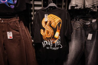 Lviv, Ukrayna - 13 Mart 2025: Siyah Grafik Para Konuşmaları ve Altın Fısıltılar tişörtü Bir dolar işareti tasarımı, bir elbise sergisinde asılı