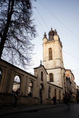 Lviv, Ukrayna - 31 Mart 2025: Mimari detaylarla donatılmış şık kilise kulesi, Lviv, Ukrayna yakınlarındaki insanlarla sokak manzarası çerçeveli. 