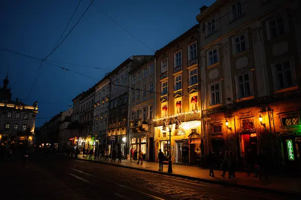 Lviv, Ukrayna - 31 Mart 2025: Aydınlatılmış binalar ve Ukrayna 'nın Lviv kentindeki eski bir şehir caddesinde insanların sergilendiği canlı bir gece sahnesi. 
