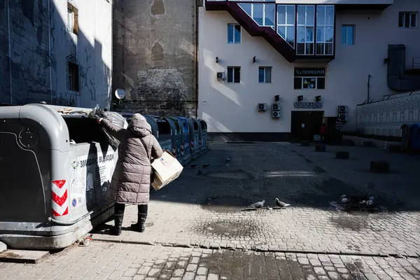 Lviv, Ukrayna - Nisan 06, 2025: Ceket giyen yaşlı bir kadın dışarıda büyük geri dönüşüm kutularına mukavva bırakıyor.