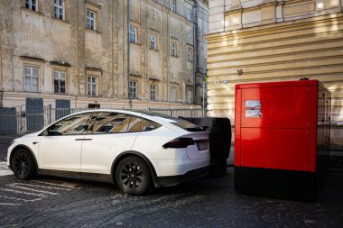 Lviv, Ukrayna - 07 Nisan 2025: Beyaz elektrikli Tesla Model X aracı kırmızı bir kutunun yanına park edilmiş eski bir binanın arka planına karşı