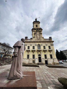 Buchach, Ukrayna - 08 Nisan 2025: Taras Hryhorovych Shevchenko 'nun bulutlu bir gökyüzüne karşı çerçevelediği bitişik bir heykeli olan süslü tarihi bir bina. Buchach Belediye Binası Kulesi. 