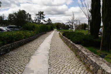Parlak bulutlu gökyüzünün altında yeşillik ve ağaçlarla kaplı manzaralı bir taş patika..