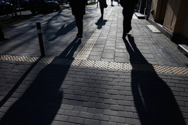 Şehir caddesinde parlak güneş ışığı altında yürüyen insanların siluetleri.