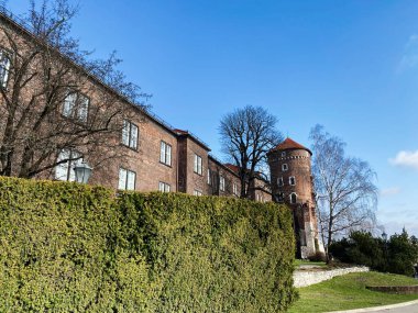 Taştan kulesi olan kırmızı tuğla mimari, açık mavi gökyüzünün altında yemyeşil çalılar..