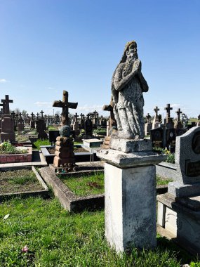 Khorostkiv, Ukrayna - 23 Nisan 2025: Yeşil çimlerin üzerinde yıpranmış mezar taşlarının arasında taş bir heykel bulunan tarihi bir mezarlık.