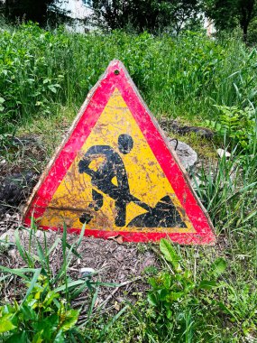 Eski üçgen yol kenarı tabelasında aşırı büyümüş çimenler ve bitki örtüsüyle çevrili inşaat uyarısı görünüyor.