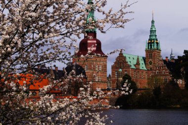 Hillerod, Danimarka - 21 Nisan 2025: Güzel bahar çiçekleri sakin bir nehir kıyısındaki görkemli bir Danimarka Frederiksborg şatosunu çerçeveler. Hillerd, Danimarka. 