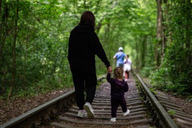Yoğun yeşil ağaçların arasında tren raylarında yürürken bir kadın çocuğun elini tutuyor.