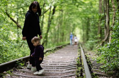 Bir anne, küçük çocuğuyla birlikte ormandaki geniş bir patika boyunca yürür..