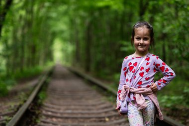 Kalp desenli bir gömlek giyen neşeli bir çocuk manzaralı bir demiryolu yolunda duruyor..
