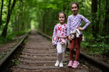 İki neşeli çocuk, yemyeşil ormanlarla çevrili eski tren raylarında duruyor.