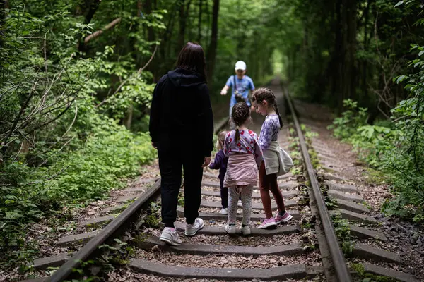 Çevresi çocuklarla çevrili bir orman raylarında yürüyen bir aile.