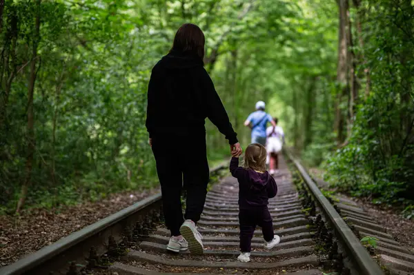 Yoğun yeşil ağaçların arasında tren raylarında yürürken bir kadın çocuğun elini tutuyor.