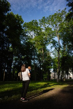Uzun yeşil ağaçlarla çevrili güneşli bir parkta çocuğunu taşıyan bir kadın..