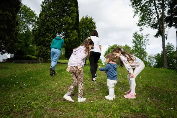 Çocuklar ve yetişkinler eğlenceli etkileşimlerle dolu yemyeşil bir parkta birlikte yürüyorlar.