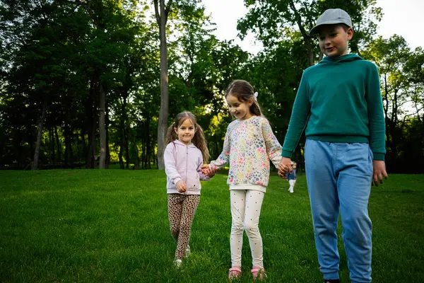 Yeşil çimenli bir parkta yürüyen ve el ele tutuşan üç çocuk.
